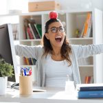 Happy young business woman having fun and holding red apple over her head in the office.